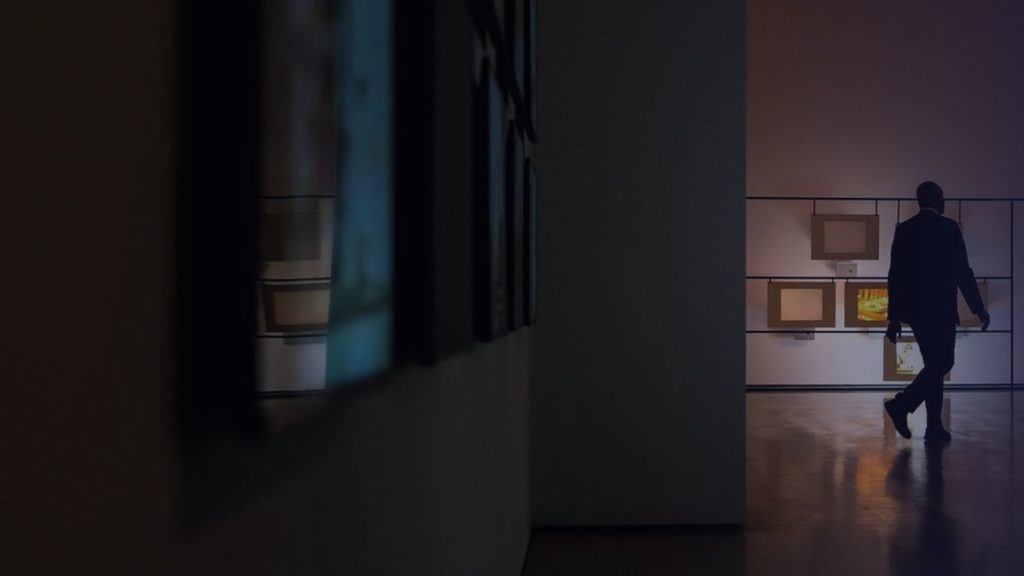 A visitor walks through a dimly lit gallery past works on the walls