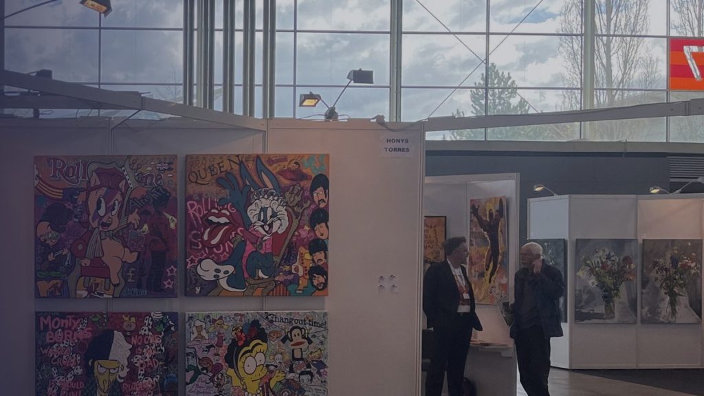 An art fair stand with bright paintings and two visitors talking